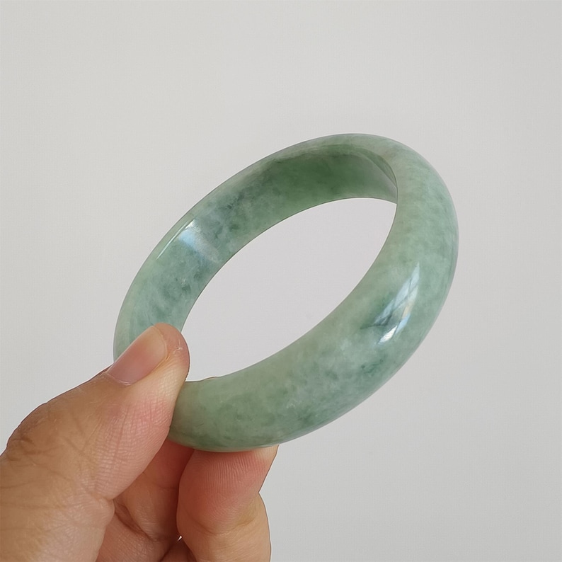May include: A pale green jade bangle. The smooth, polished bracelet has a translucent quality, with subtle colour variations. The jade band is held in a hand against a white background. The bracelet is a solid, circular band.