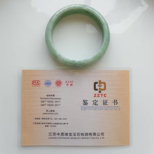 May include: A pale green jade bangle. The circular bracelet is on a white surface, next to a certificate with Chinese text and the words "IDENTIFICATION CERTIFICATE". The bracelet is a classic design.