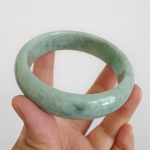 May include: A pale green jade bangle bracelet. The polished bracelet is held in a hand, highlighting its natural colour variations. The jade has a translucent quality, with subtle darker green veining.