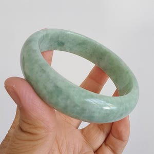 May include: A pale green jade bangle. The polished surface of the bracelet reflects light, highlighting the natural colour variations. The jade has a soft, muted green hue, with subtle darker green veining. The bracelet is a circular shape, suitable for a wrist.