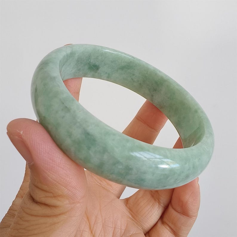 May include: A pale green jade bangle. The polished surface of the bracelet reflects light, highlighting the natural colour variations. The jade has a soft, muted green hue, with subtle darker green veining. The bracelet is a circular shape, suitable for a wrist.