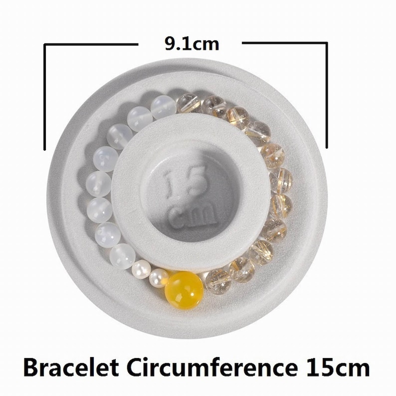 May include: A white ceramic bracelet sizing ring with a 9.1 cm diameter. The ring has a 15 cm circumference measurement printed on it. Two bracelets are displayed on the ring, one with white beads and one with gold beads.