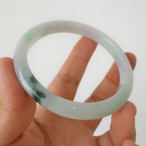 Può includere: Un braccialetto bangle in giada con una superficie liscia e lucida. Il braccialetto presenta un mix di tonalità verde chiaro, bianco e verde più scuro. La giada è traslucida, permettendo alla luce di passare attraverso. Il braccialetto è tenuto in una mano.