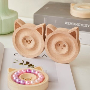 May include: Three wooden ring holders shaped like pig faces. The holders are made of light brown wood and have a small indentation in the center for rings. The holders are sitting on a white surface.