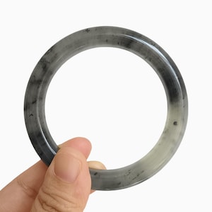 Puede incluir: Un brazalete de jade gris y blanco. El brazalete es una banda circular sólida con una superficie lisa y pulida. El color es una mezcla de gris y blanco, con vetas grises más oscuras. El brazalete está sostenido por una mano.