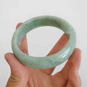 May include: A pale green jade bangle. The smooth, polished bracelet is held in a hand against a white backdrop. The jade has a translucent quality with subtle colour variations.