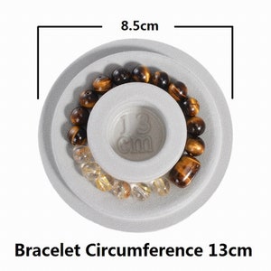 May include: A gray, round, two-tiered display stand with a brown and gold beaded bracelet on top. The stand measures 8.5 cm in diameter and the bracelet has a circumference of 13 cm.