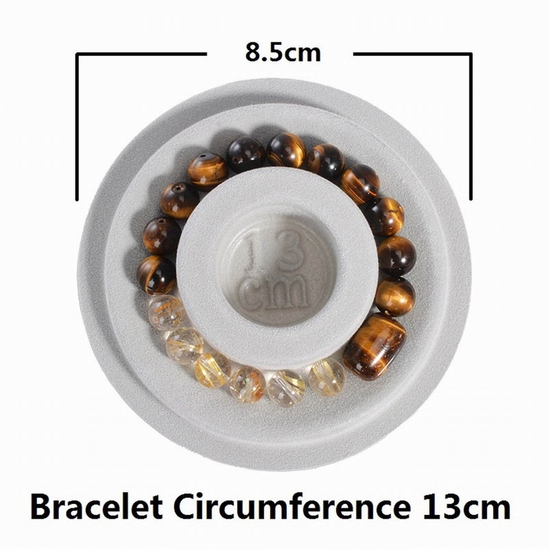 May include: A gray, round, two-tiered display stand with a brown and gold beaded bracelet on top. The stand measures 8.5 cm in diameter and the bracelet has a circumference of 13 cm.