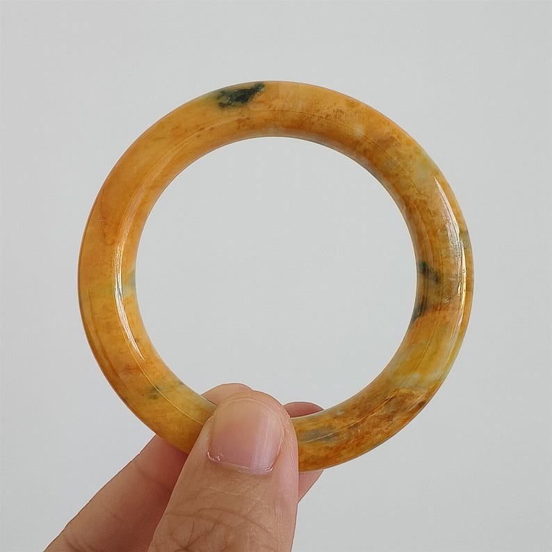 May include: A round, yellow-orange jade bangle bracelet. The bracelet has a smooth, polished surface with natural variations in color, including green and brown spots. The bangle is held against a white background.