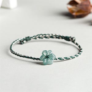 Könnte beinhalten: Ein zartes Armband mit einem hellgrünen Blumenanhänger in der Mitte. Das Armbandband ist eine Kombination aus grünen und weißen Fäden. Der Blumenanhänger hat eine hellgrüne Farbe. Das Armband ist für den täglichen Gebrauch konzipiert.