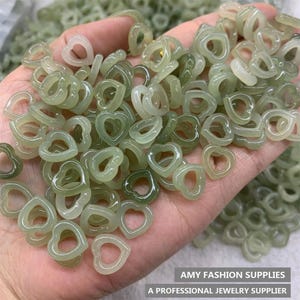 Pode incluir: Um conjunto de contas em forma de coração, translúcidas e verde-claro. As contas são pequenas e parecem ser feitas de um material liso e polido. A imagem inclui o texto "AMY FASHION SUPPLIES" e "A PROFESSIONAL JEWELRY SUPPLIER".