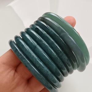 Puede incluir: Una pila de siete brazaletes redondos y lisos en varios tonos de verde bosque profundo y verde salvia translúcido. Cada brazalete presenta un acabado pulido y brillante con sutiles inclusiones minerales naturales visibles en la piedra.
