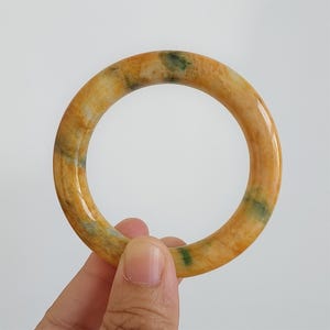 May include: A round jade bangle bracelet. The bracelet is a mix of yellow, orange, and green hues. The smooth, polished surface reflects light, highlighting the natural variations in color. A piece of jewelry.
