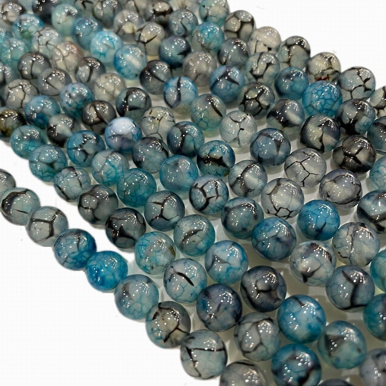 May include: Close-up of a collection of round, blue and grey beads. The beads have a cracked, web-like pattern. The beads are strung together, suggesting they are for jewellery making or crafts.