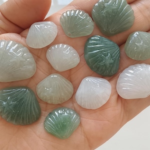 Peut inclure: Une collection de pendentifs en forme de coquillages sculptés, de différentes nuances de vert et de blanc. Les pendentifs sont tenus dans une main, mettant en valeur leurs détails complexes et leurs variations naturelles. Les coquillages sont lisses et polis.