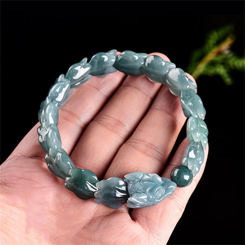 Puede incluir: Un brazalete de jade con un dise&ntilde;o floral. El brazalete est&aacute; compuesto por m&uacute;ltiples piezas de jade talladas en tonos verdes y blancos. El brazalete se muestra sobre un fondo oscuro.