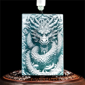 Puede incluir: Colgante rectangular de color turquesa con un detallado tallado de dragón. El dragón está representado con detalles intrincados y acentos blancos. El colgante cuelga de un cordón negro. El diseño general es un dragón.