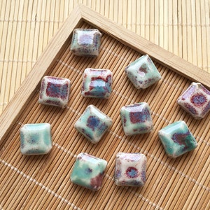 Puede incluir: Doce azulejos cuadrados de cerámica con un esmalte moteado en tonos de verde, marrón y blanco. Los azulejos están dispuestos en un patrón de cuadrícula sobre una estera de bambú.