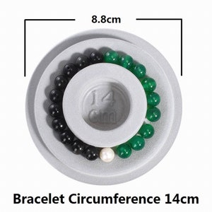 May include: A white measuring tool with a 14 cm diameter circle for measuring bracelet circumference. The tool has a black and green beaded bracelet on it.