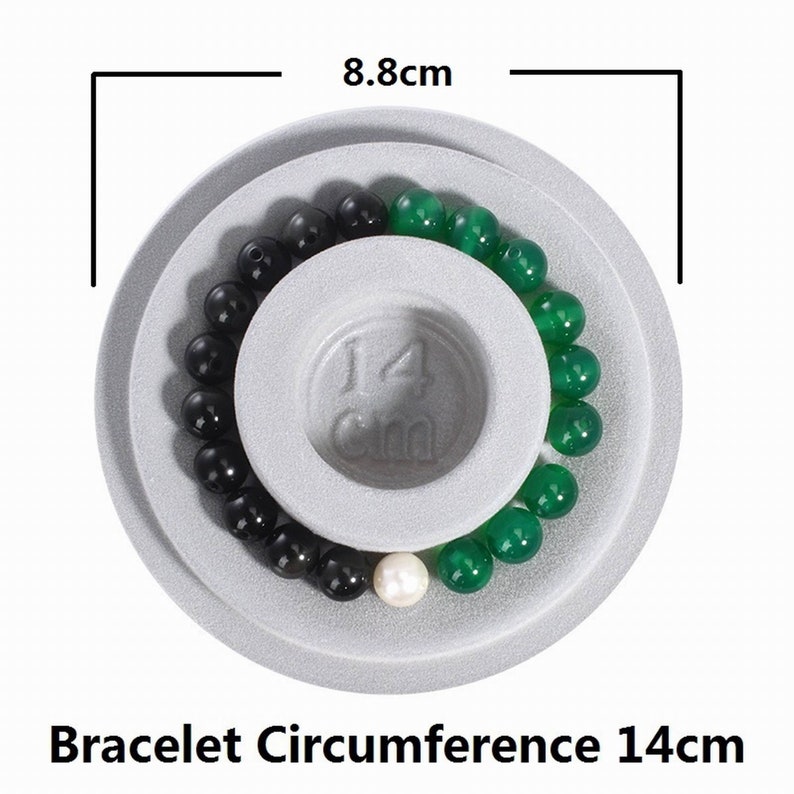 May include: A white measuring tool with a 14 cm diameter circle for measuring bracelet circumference. The tool has a black and green beaded bracelet on it.
