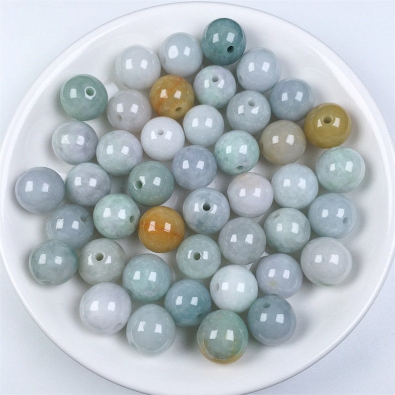 May include: A collection of round, smooth jade beads in various shades of green, white, and yellow. The beads are arranged in a bowl.