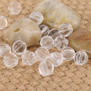 May include: A collection of clear, faceted, round glass beads. The beads are scattered on a brown burlap surface with two light brown stones.