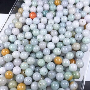 May include: A large collection of round, smooth jade beads in various shades of green, white, and yellow. The beads are arranged in a clear container.