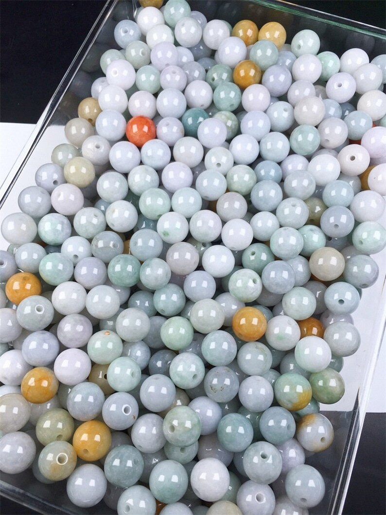 May include: A large collection of round, smooth jade beads in various shades of green, white, and yellow. The beads are arranged in a clear container.