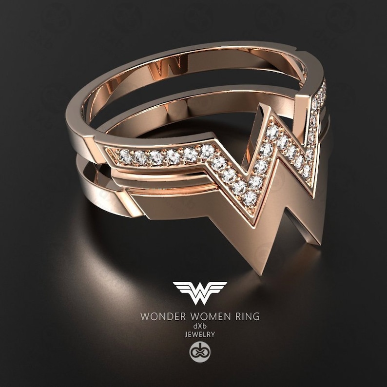 Może przedstawiać: Pierścionek z r&oacute;żowego złota z logo Wonder Woman, inkrustowany małymi, błyszczącymi kamieniami. Pierścionek ma podw&oacute;jną opaskę z kultowym symbolem "W". Biżuteria.