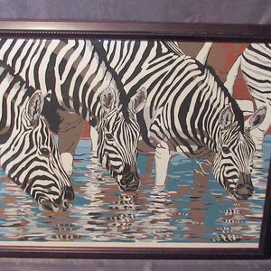 Könnte beinhalten: Gerahmtes Gemälde mit Zebras, die Wasser trinken. Das Kunstwerk zeigt vier Zebras mit schwarz-weißen Streifen, die ihre Köpfe senken, um aus dem Wasser zu trinken. Das Wasser spiegelt die Zebras und den Himmel wider und erzeugt eine ruhige Szene.