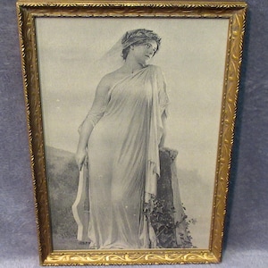 May include: A framed black and white print depicting a figure in a draped gown and a laurel wreath headpiece, leaning against a stone structure. The artwork is enclosed in a rectangular gold-toned frame with a textured, ornate border design.