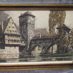 May include: A framed print depicting a historic European town scene with a stone bridge, a tall tower, and timber-framed buildings situated along a riverbank. The artwork features muted earth tones, including browns, grays, and greens, enclosed in a simple wooden frame.