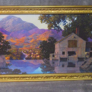 May include: A rectangular landscape print in a decorative gold frame. The artwork depicts a light-colored wooden building situated by a calm body of water, with large trees and purple-toned mountains in the background under a clear sky.