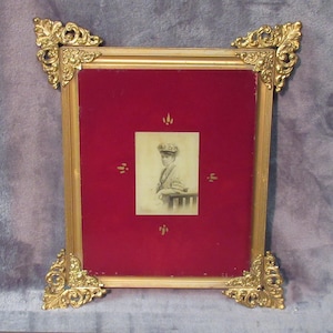 May include: A vintage gold-toned picture frame with ornate corner details. The frame holds a small black and white portrait set against a red velvet background. The frame is square and has a classic, antique aesthetic.