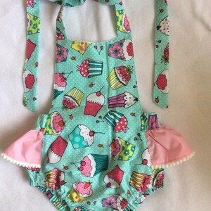 May include: A turquoise baby onesie with a cupcake print and pink ruffle trim. The onesie has a halter neck style and ties at the back.