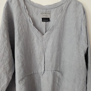 May include: A light blue linen top with a V-neck and long sleeves. The top has a relaxed fit and a raw edge detail along the neckline. The tag reads "breathe clothing".