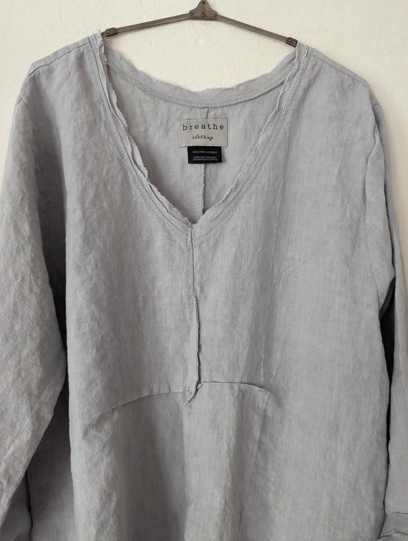 May include: A light blue linen top with a V-neck and long sleeves. The top has a relaxed fit and a raw edge detail along the neckline. The tag reads "breathe clothing".