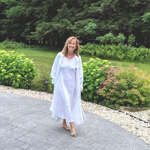 K&ouml;nnte beinhalten: Eine Person tr&auml;gt ein wei&szlig;es Leinenkleid und einen passenden langen Mantel und geht barfu&szlig; auf einer Steinterrasse. Der Hintergrund zeigt &uuml;ppiges gr&uuml;nes Laub und B&auml;ume, die eine ruhige Umgebung im Freien schaffen.