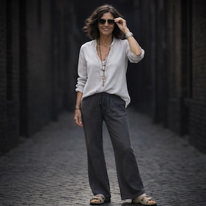 May include: A woman wearing a light gray long-sleeve shirt, dark gray pants, and sandals stands in a cobblestone alley. She wears sunglasses and a necklace. The outfit is casual and comfortable.