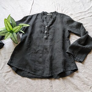 May include: A dark gray linen blouse with long, flared sleeves and a ruffled neckline. The blouse has a button-up front and a relaxed fit. A potted plant with green and white leaves is placed near the blouse.