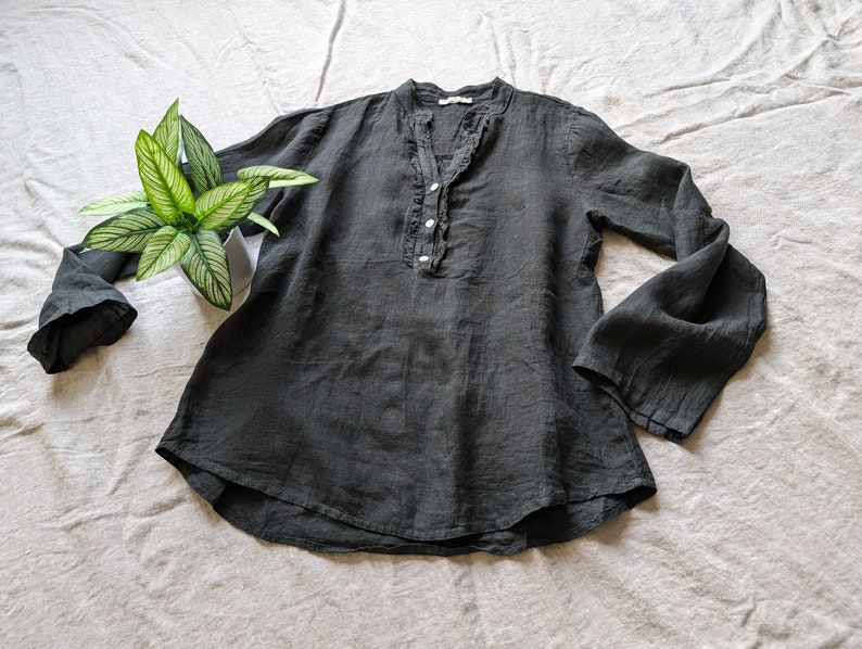 May include: A dark gray linen blouse with long, flared sleeves and a ruffled neckline. The blouse has a button-up front and a relaxed fit. A potted plant with green and white leaves is placed near the blouse.