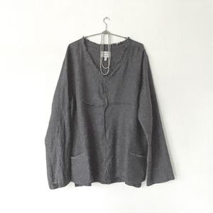 May include: A dark gray long-sleeve shirt with a V-neck and two front pockets. The shirt is hanging on a metal hanger against a white background. A silver necklace is draped on the shirt.