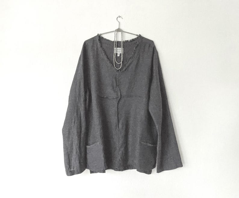 May include: A dark gray long-sleeve shirt with a V-neck and two front pockets. The shirt is hanging on a metal hanger against a white background. A silver necklace is draped on the shirt.