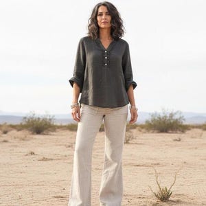 May include: A woman wearing a dark gray linen blouse with a V-neck and button details, paired with light beige linen pants. The outfit is suitable for casual wear. The background is a desert landscape.