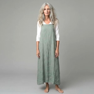 May include: A sage green linen apron dress with wooden button fastenings on the straps, worn over a white long-sleeved top. The model is barefoot against a grey backdrop. The dress has a relaxed, flowing silhouette, suitable for layering.