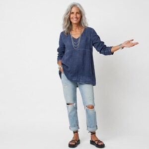 May include: A woman wearing a dark blue V-neck tunic, light blue ripped jeans, and black sandals. The tunic has long sleeves and a relaxed fit. The jeans are cuffed at the ankles. She is wearing a silver necklace and a watch.