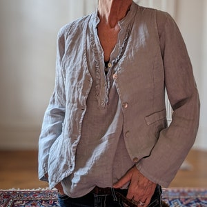 May include: A light gray linen jacket with a matching shirt underneath. The jacket has a classic blazer design with buttons and pockets. The shirt has a ruffled neckline. The outfit is paired with dark blue jeans.