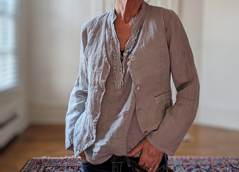 May include: A light gray linen jacket with a matching shirt underneath. The jacket has a classic blazer design with buttons and pockets. The shirt has a ruffled neckline. The outfit is paired with dark blue jeans.
