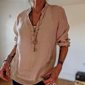 May include: A light beige linen blouse with a ruffled collar and long sleeves. The blouse has a button-down front and is paired with a long necklace featuring a key pendant. The sleeves are rolled up, and the blouse has a relaxed fit.