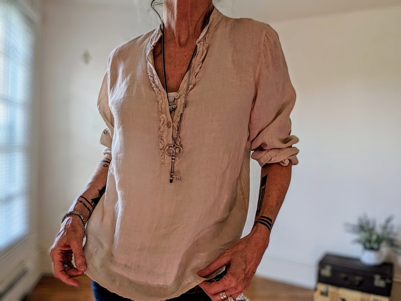 May include: A light beige linen blouse with a ruffled collar and long sleeves. The blouse has a button-down front and is paired with a long necklace featuring a key pendant. The sleeves are rolled up, and the blouse has a relaxed fit.